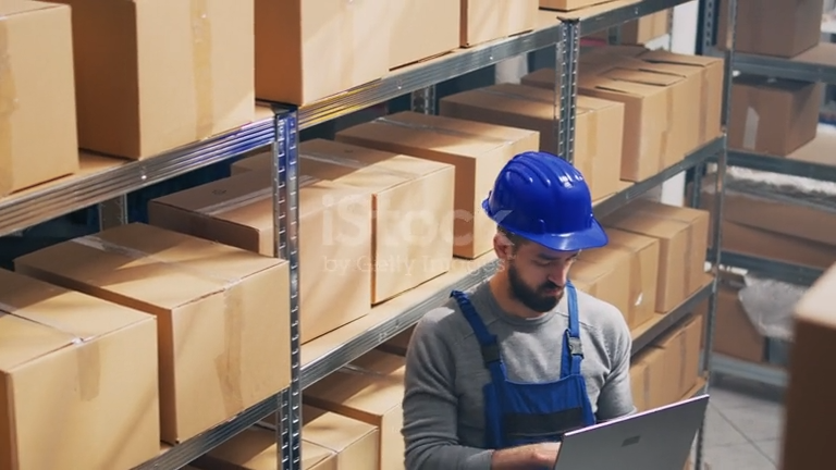 Agent en entrepôt avec casque bleu, travaillant sur un ordinateur portable parmi des rayonnages de cartons. smic horaire nuit à considérer dans le calcul et la majoration.